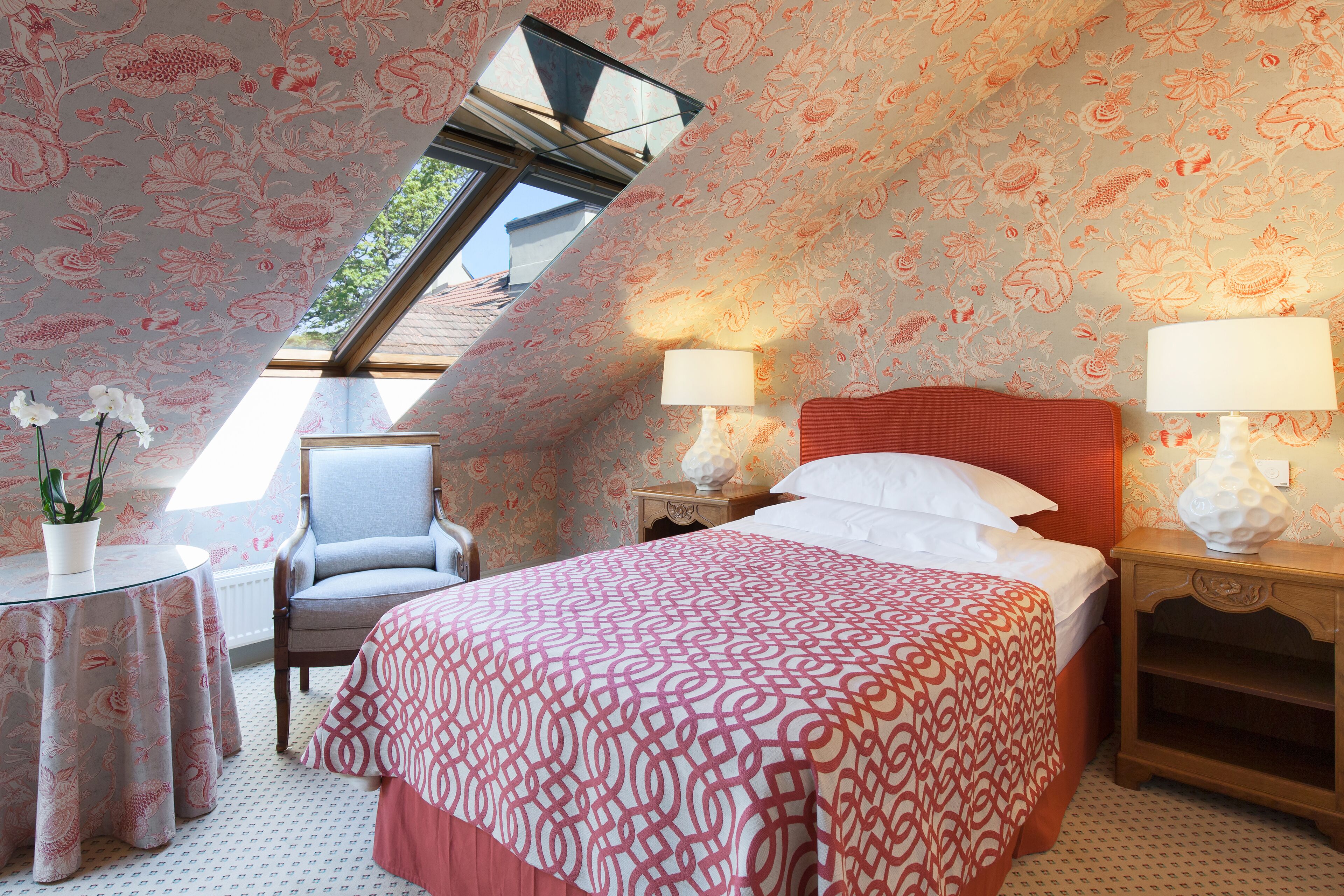 Ein Schlafzimmer mit schrägem Dach, blumigem Wandpapier, einem Bett mit rotem Kopfteil, zwei Nachttischen mit Lampen, einem Sessel und einem kleinen Tisch mit einer Pflanze.