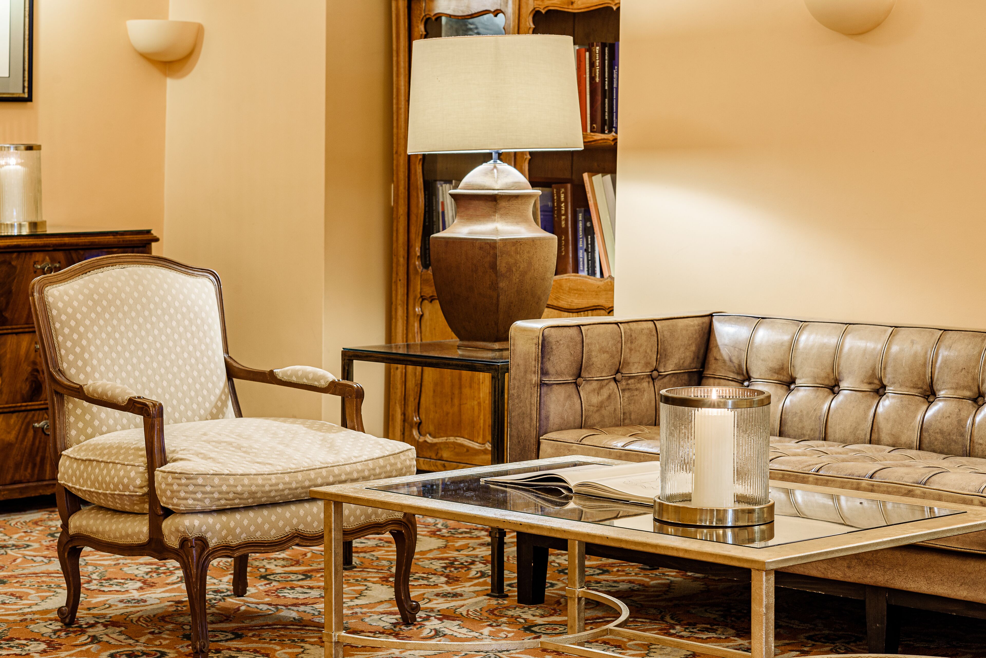 Una sala de estar con un sillón estampado, un sofá de cuero, una mesa de centro de vidrio, una lámpara y una estantería.