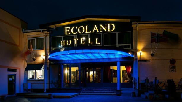 The exterior of the Ecoland Hotel with a prominent illuminated sign, a covered entrance, and a flag on the right side. - Ecoland Hotel (Tallinn)