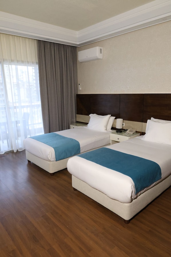 A hotel room with two beds, a wooden floor, a window with curtains, and a wall-mounted air conditioner.