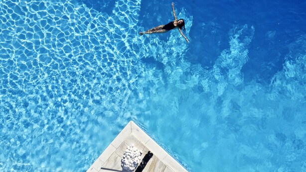 A person swimming in a clear blue pool with a tiled deck area visible below.