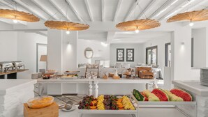 A breakfast buffet with various fruits and pastries, white furniture, and woven ceiling lights.