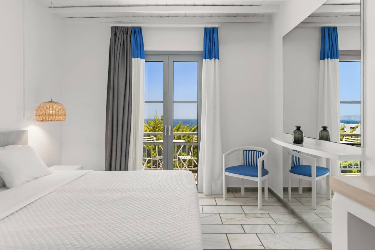 Habitación de hotel con cama, escritorio y sillas, vista al exterior y balcón con sillas.