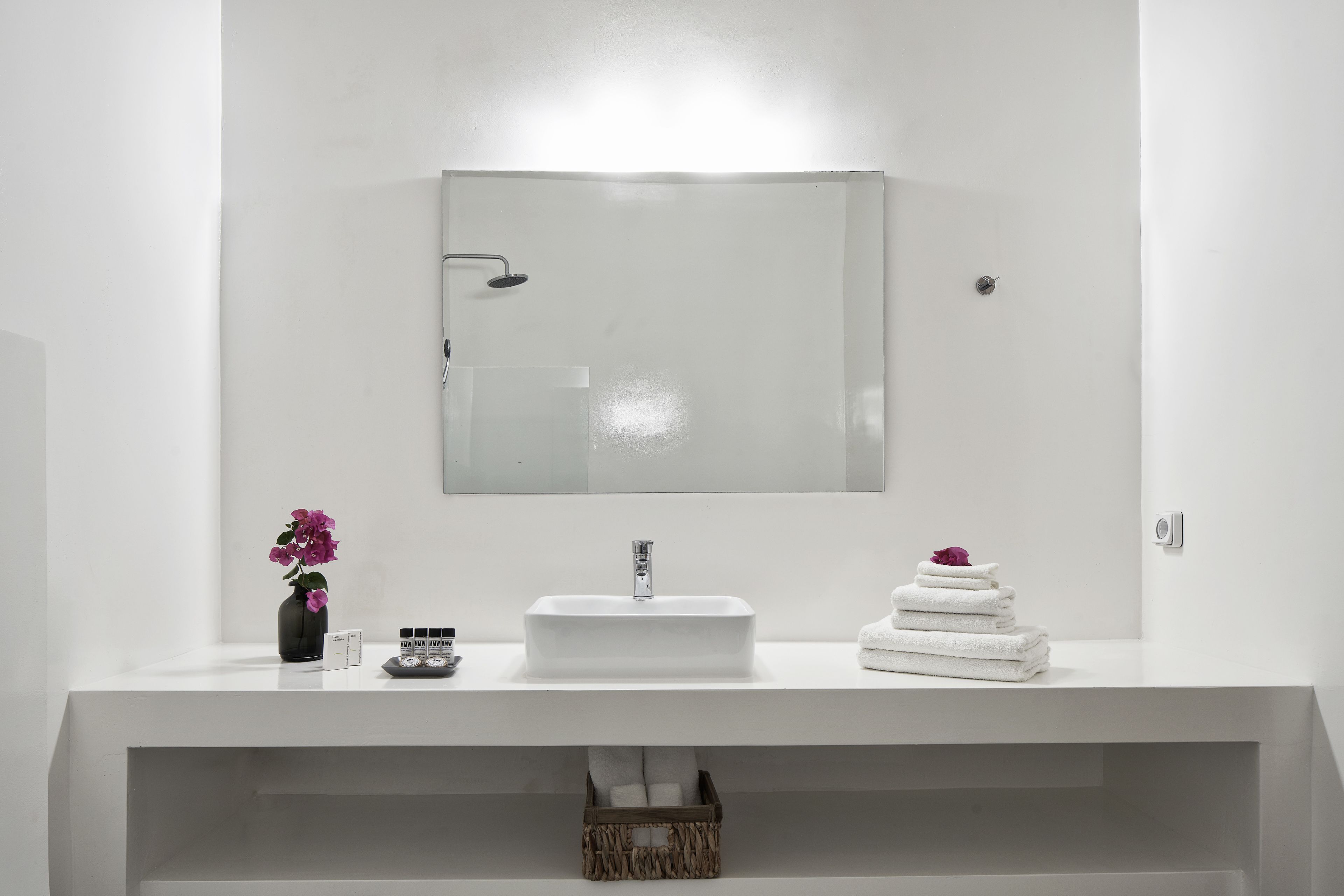 Un baño moderno con un tocador blanco, un espejo rectangular, un lavamanos empotrado y un pequeño jarrón con flores.