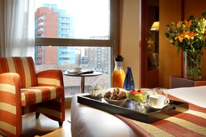 A hotel room with a bed, a striped armchair, a tray with breakfast items, a vase with flowers, and a city view through the window.