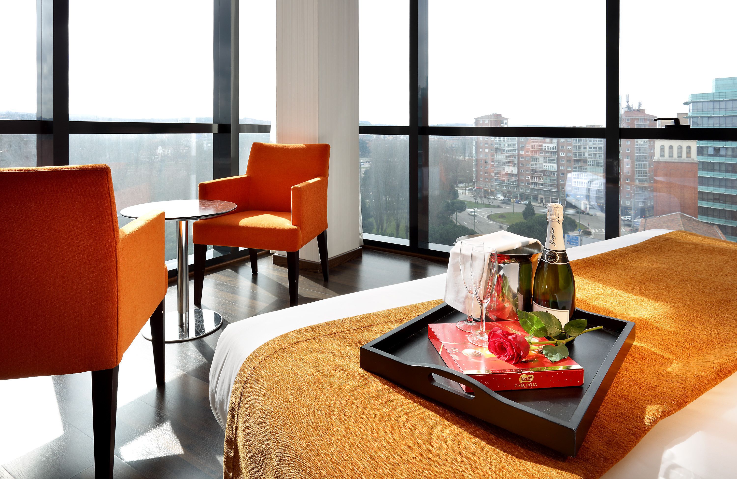 A hotel room with a large window, a bed with a tray of items, a table, and two orange chairs.