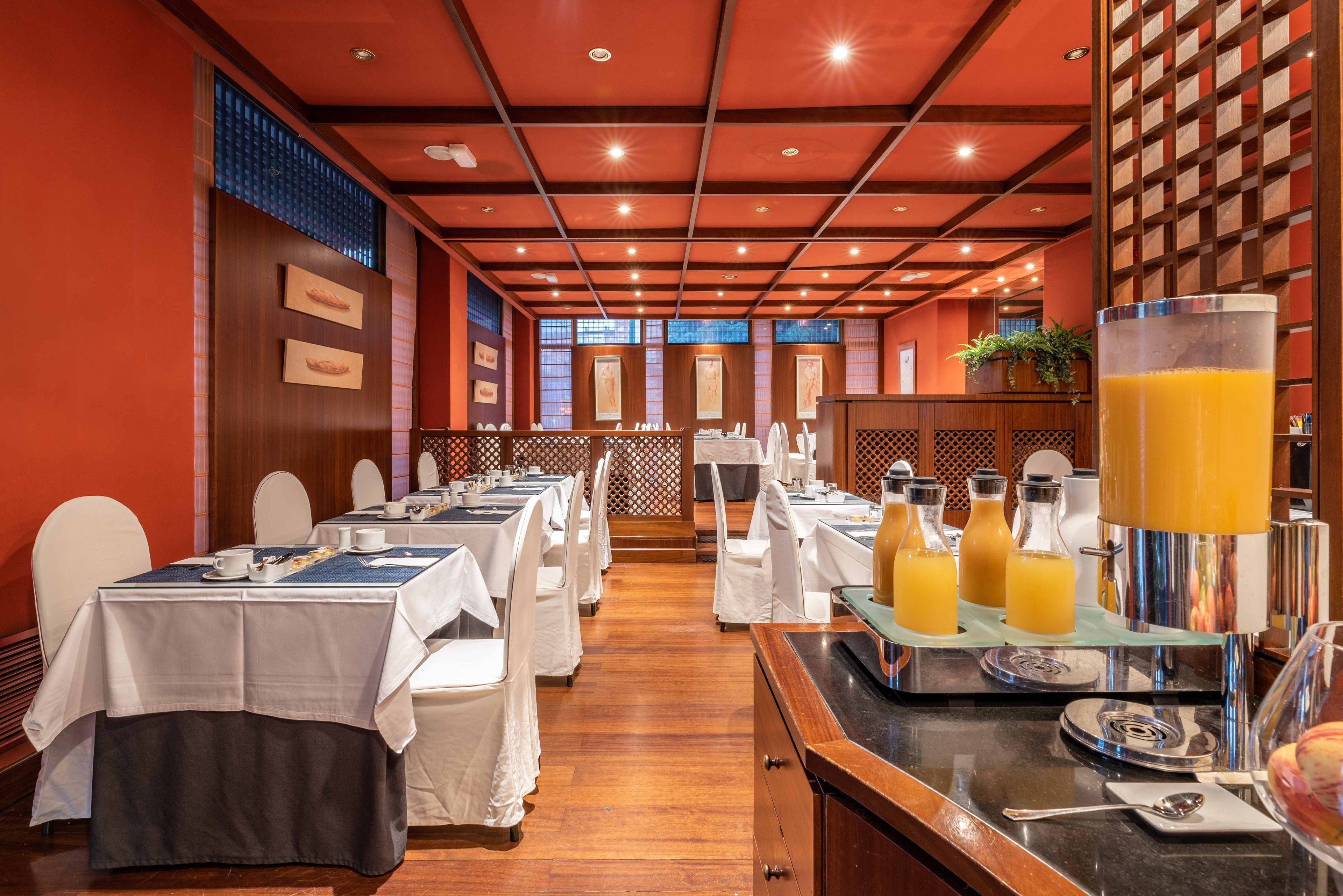 Un restaurant avec de nombreuses tables dressées pour le service, un bar doté d’un grand distributeur de jus de fruits et une vue sur l’extérieur grâce à de grandes fenêtres.