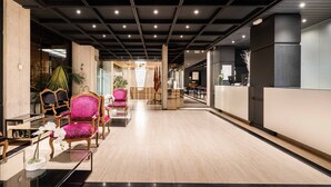 A modern lobby with a wooden floor, a reception desk, and several pink chairs.