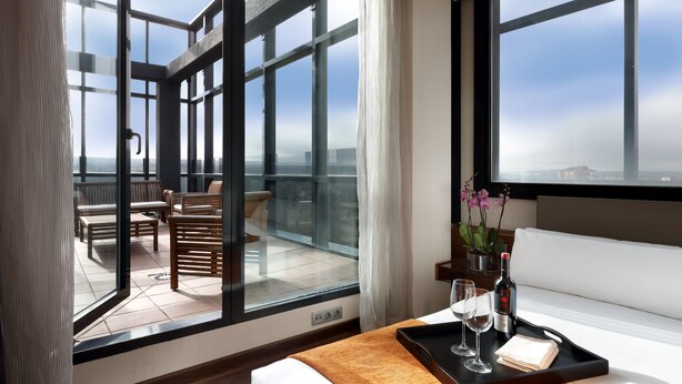 A hotel room with a large window, a balcony with seating, a bed, and a tray with a wine bottle and glasses.