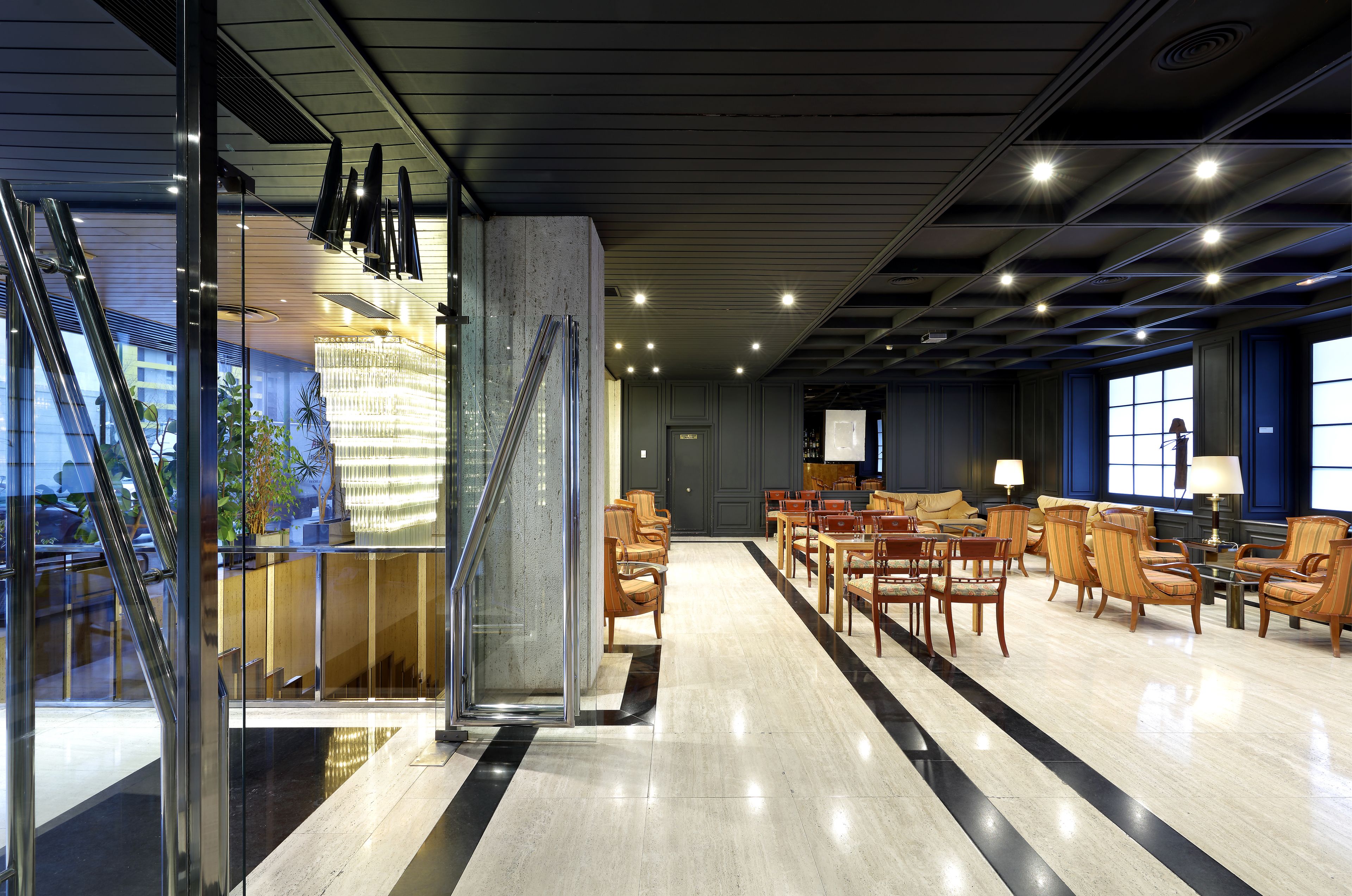 Een moderne lobby met een glazen wand, houten stoelen en een kroonluchter.