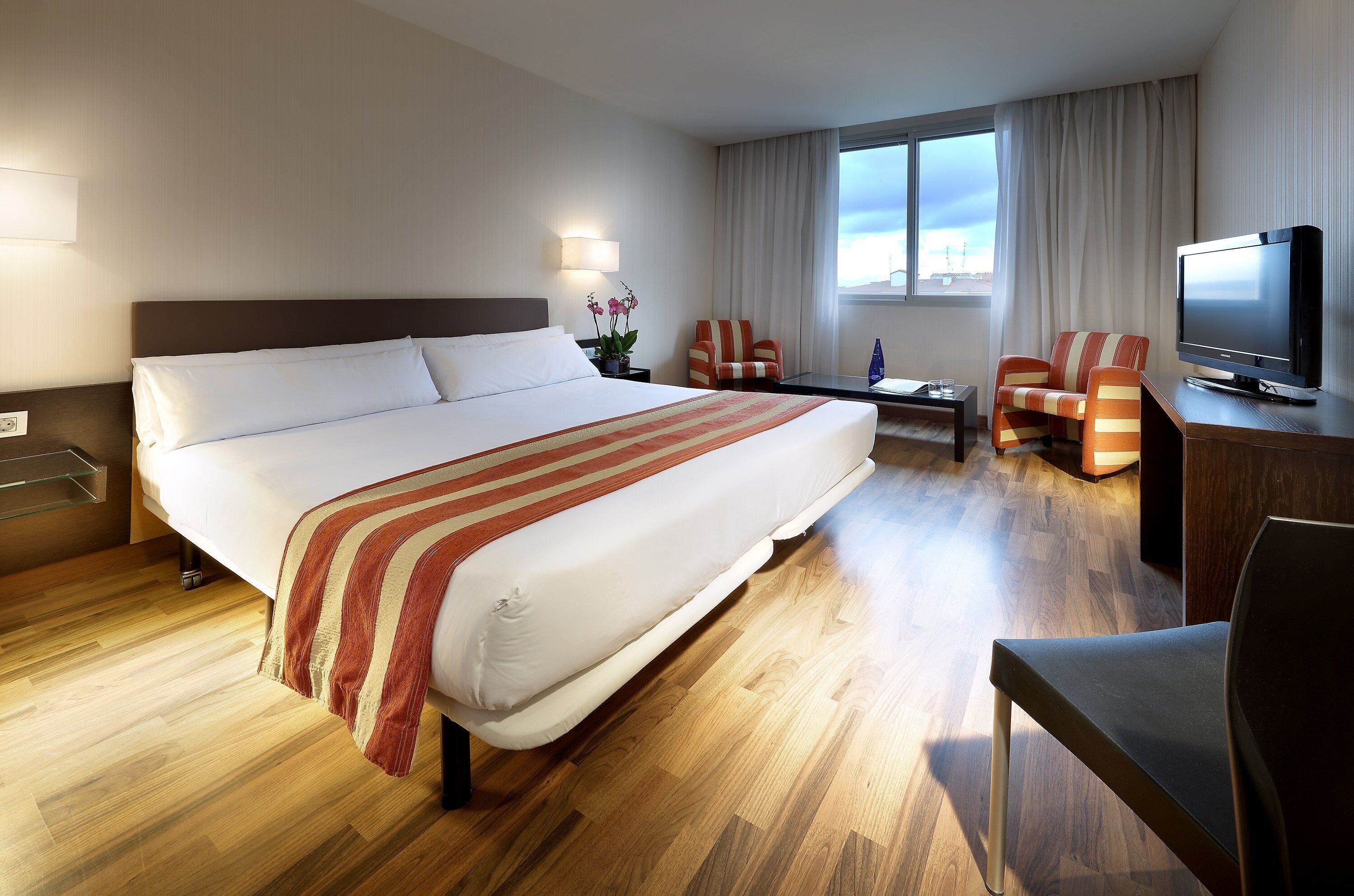 A hotel room with a large bed, a desk with a TV, two chairs, a small table, and a window with curtains.