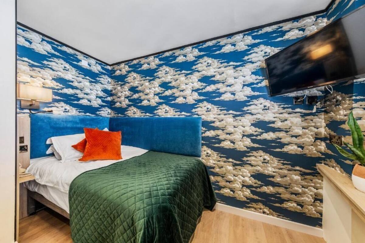 Une chambre avec un mur décoré d’un motif de nuages bleus et blancs, une couvre-lit verte et un téléviseur fixé au mur.