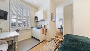 A compact room with a kitchenette, a dining table with chairs, a bed with a green quilt, and a window with blinds.
