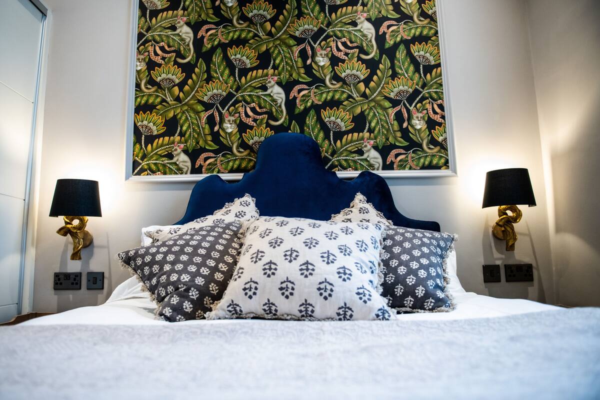 Un lit bien fait, avec une tête de lit bleue, plusieurs coussins à motifs et deux lampes fixées au mur.