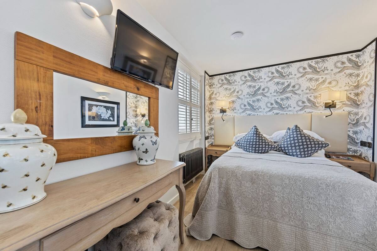 Une chambre à coucher avec une tête de lit en bois, un téléviseur à écran plat fixé au-dessus du lit, une table de chevet agrémentée d’objets décoratifs et un papier peint à motifs.