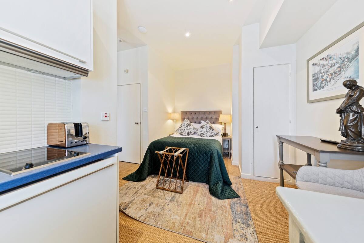 Une chambre d’hôtel compacte avec une kitchenette, un lit recouvert d’une courtepointe verte matelassée, un petit tabouret en bois, une statue posée sur une table et un tableau encadré accroché au mur.