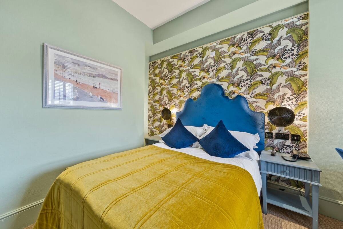 Une chambre avec un lit, une table de chevet, un ventilateur fixé au mur et un tableau encadré.