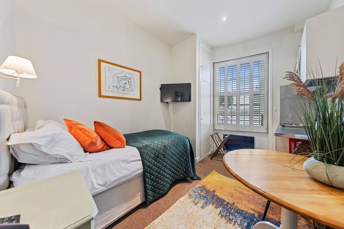 Une chambre à coucher de taille réduite, comprenant un lit, un bureau, une chaise, un téléviseur et une fenêtre avec des stores.