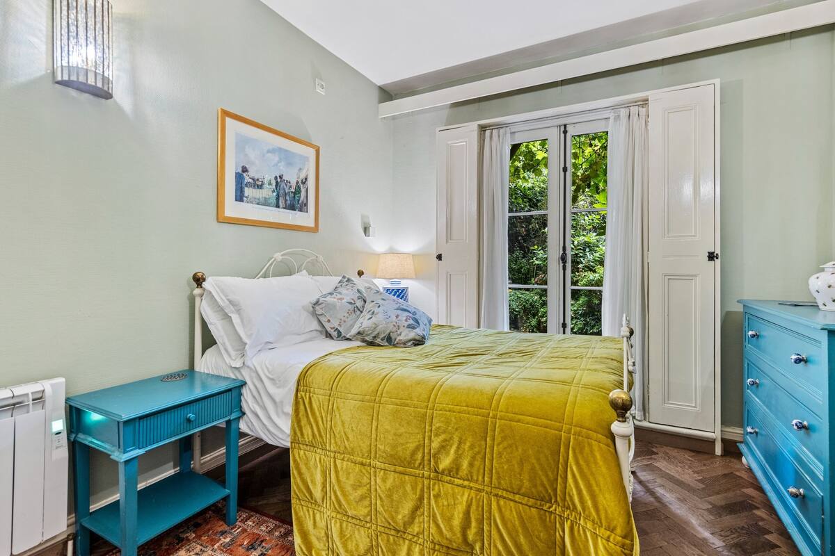 Une chambre à coucher comprenant un lit, une table de chevet, une commode, une fenêtre avec des rideaux et un tableau encadré accroché au mur.