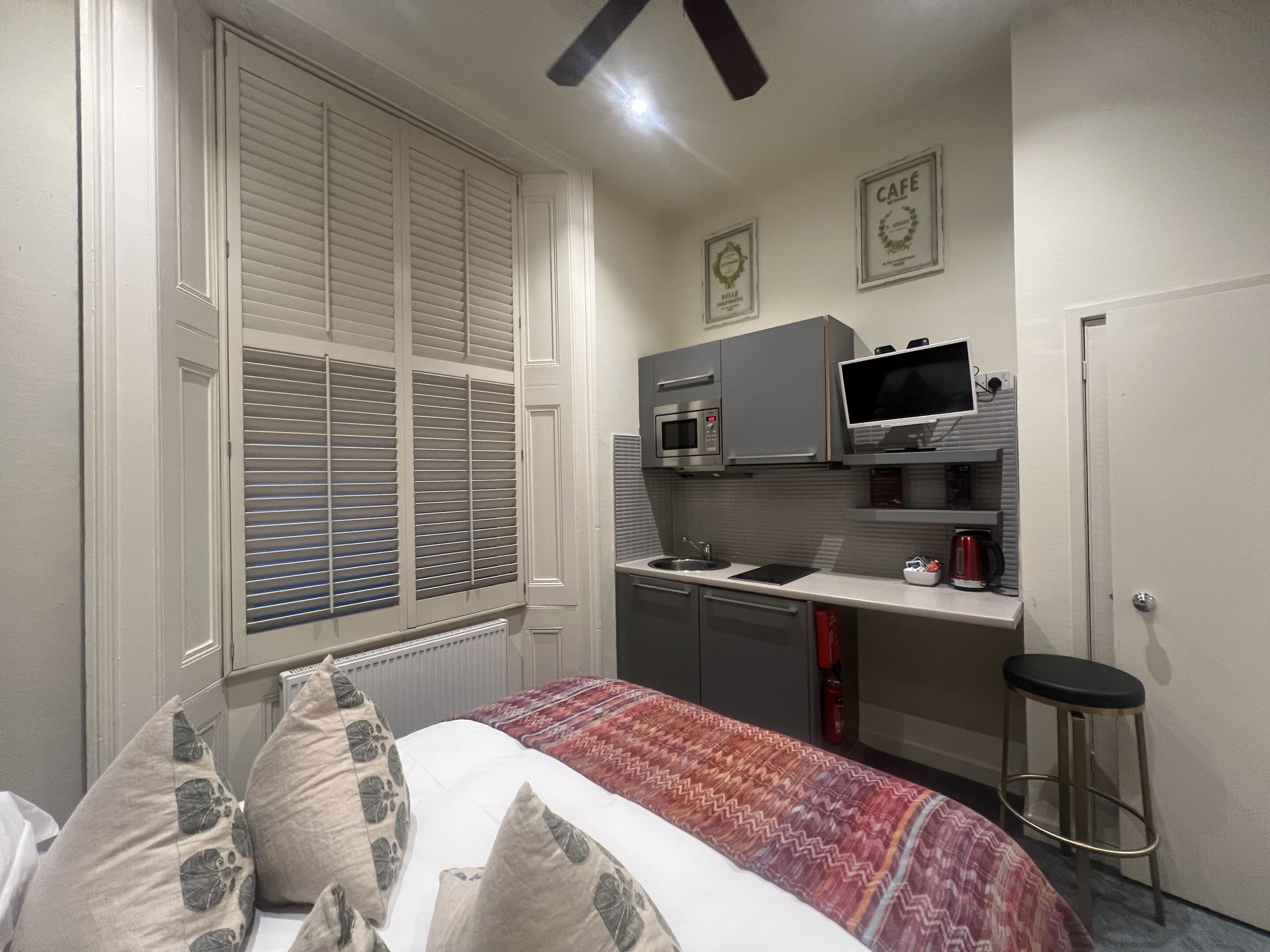 A compact bedroom with a bed, a window with blinds, a kitchenette with a microwave, a TV, and a small stool.