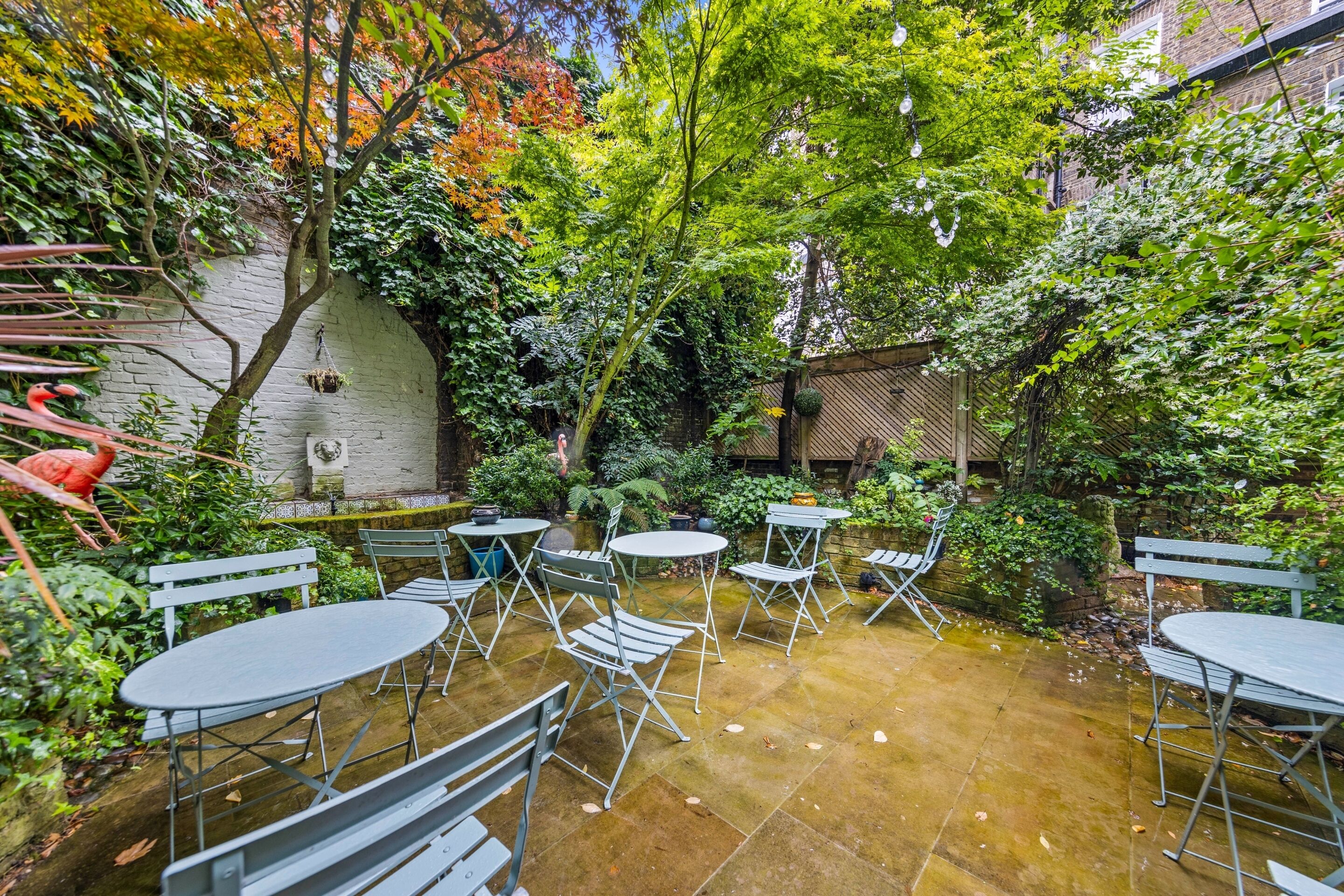 An outdoor patio with round tables and metal chairs, surrounded by lush greenery and trees.