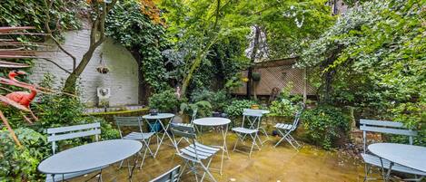 An outdoor patio with round tables and metal chairs, surrounded by lush greenery and trees.