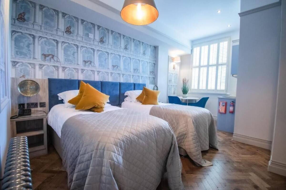 Une chambre d’hôtel avec deux lits, une tête de lit bleue, une fresque murale et une fenêtre avec des stores.