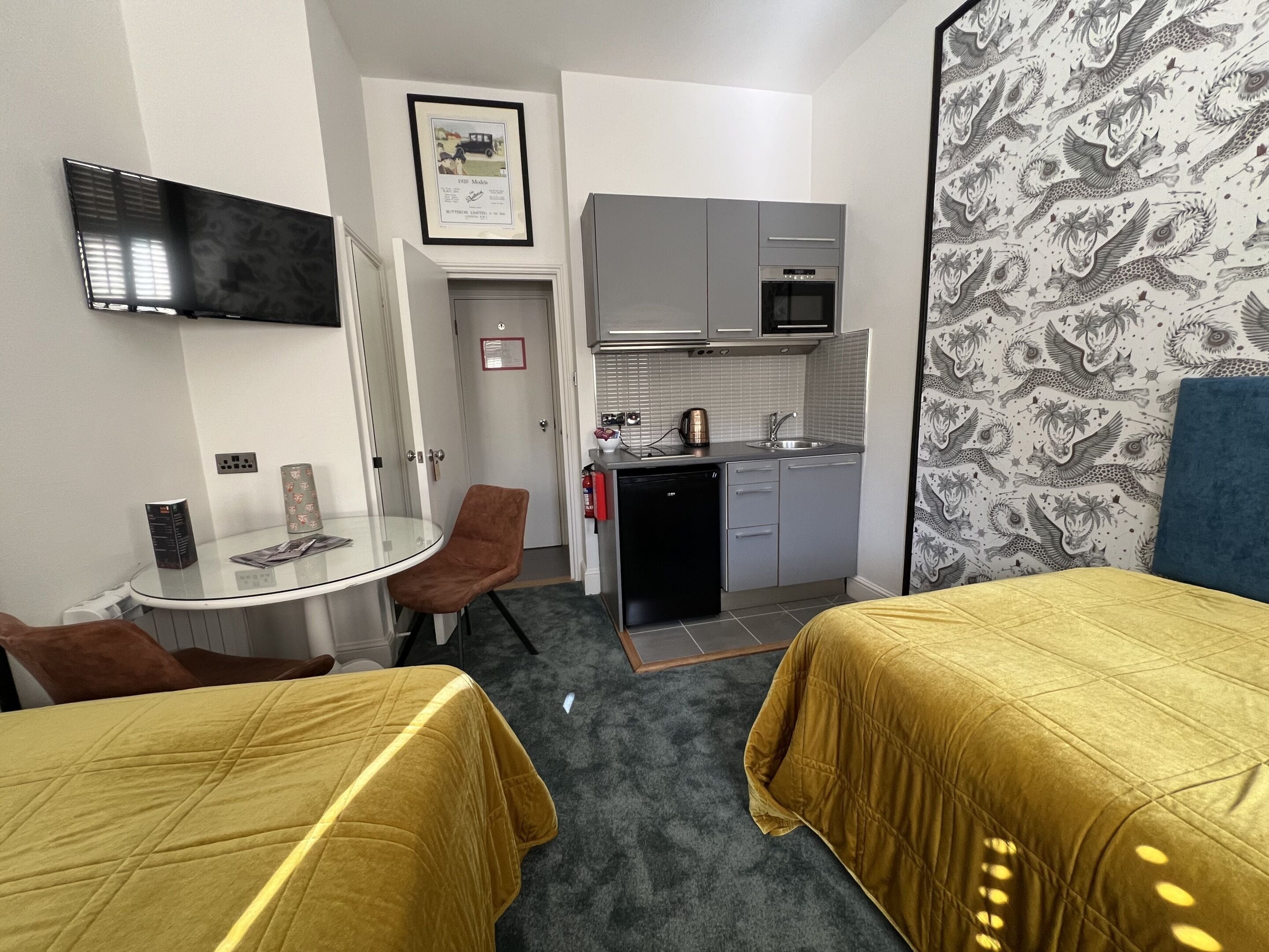 A compact hotel room with a kitchenette, a small dining area, and a bed with a yellow bedspread.