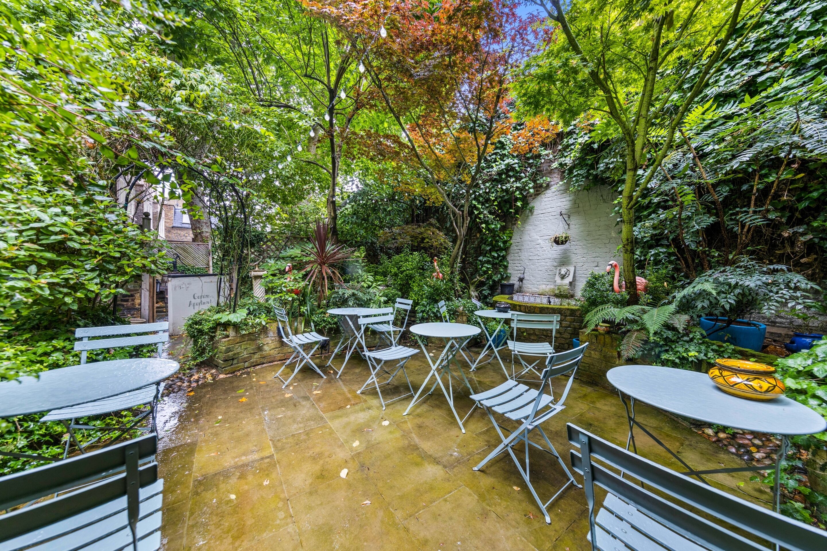 An outdoor patio with metal tables and chairs, surrounded by lush greenery and a stone wall.