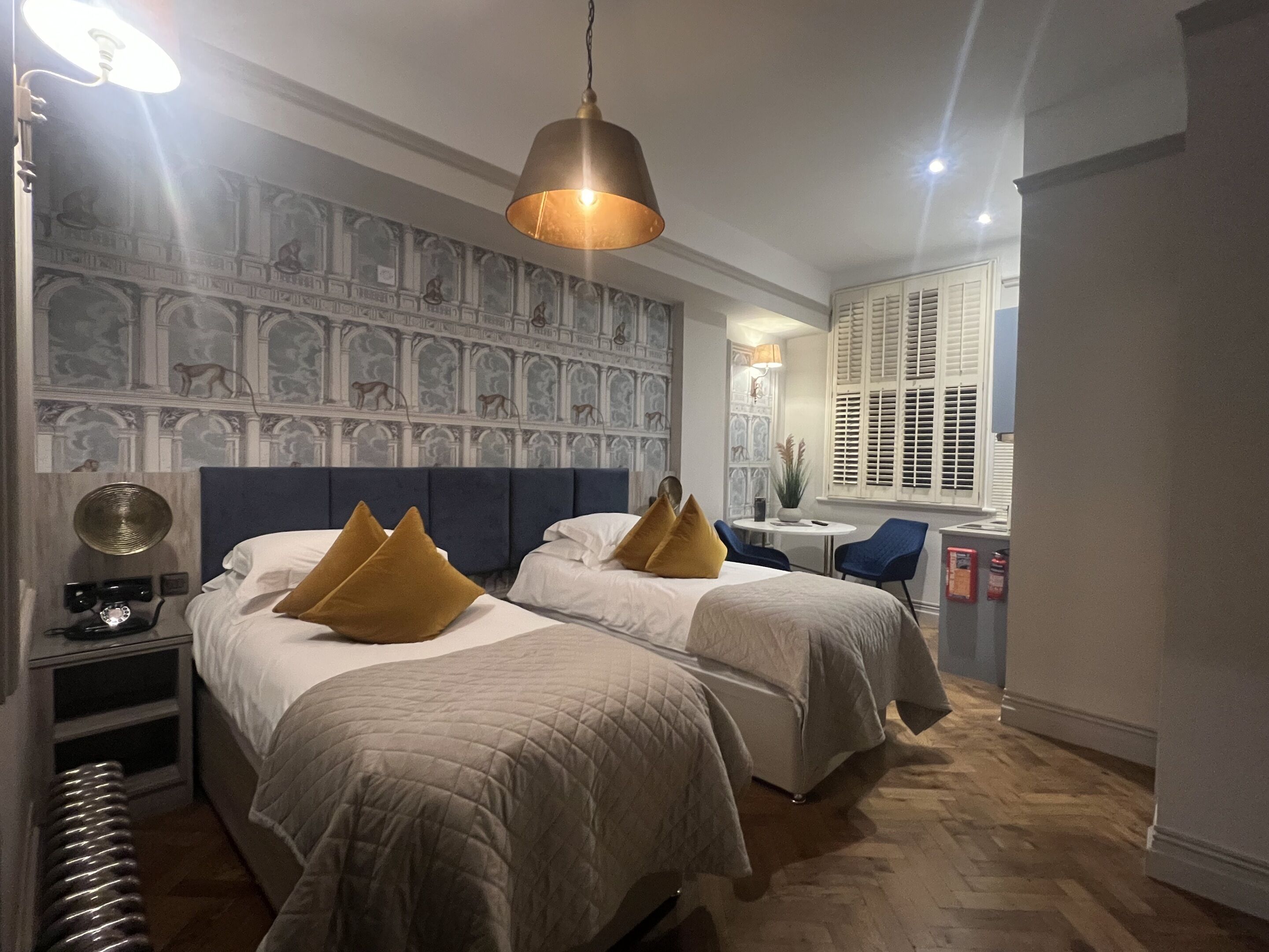 A hotel room with two beds, a large mural on the wall, a round table, and a window with shutters.