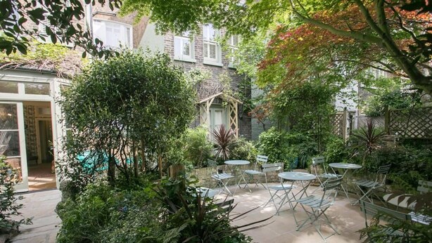 A courtyard with a glass door entrance, surrounded by lush greenery and a small wooden structure. - Vancouver Studios (London)