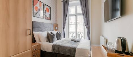 Un dormitorio con una cama, un armario de madera, una mesita de noche con hervidor y un televisor fijado a la pared.