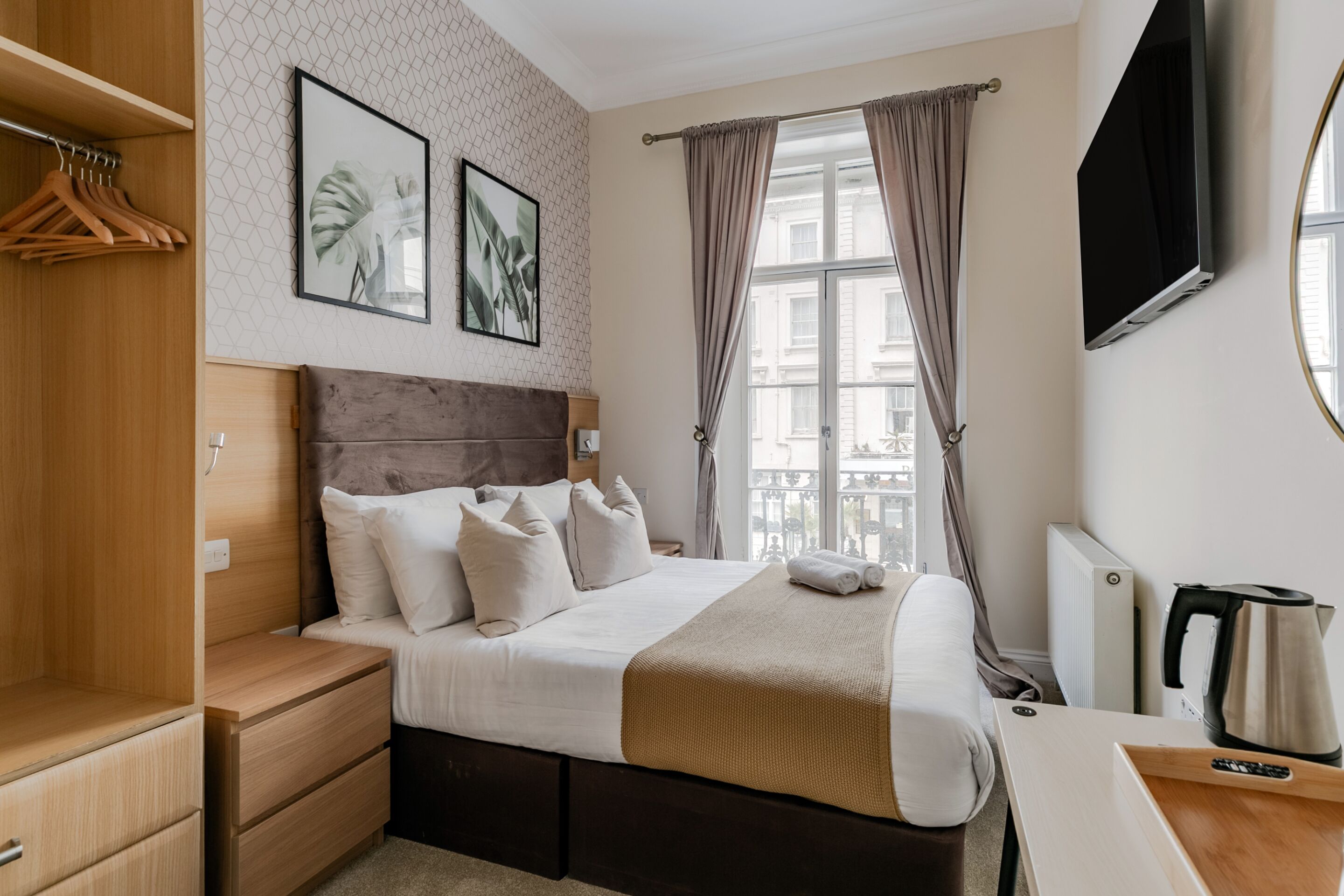 A hotel room with a bed, wooden wardrobe, two framed pictures, a window with curtains, a radiator, and a kettle on a table.