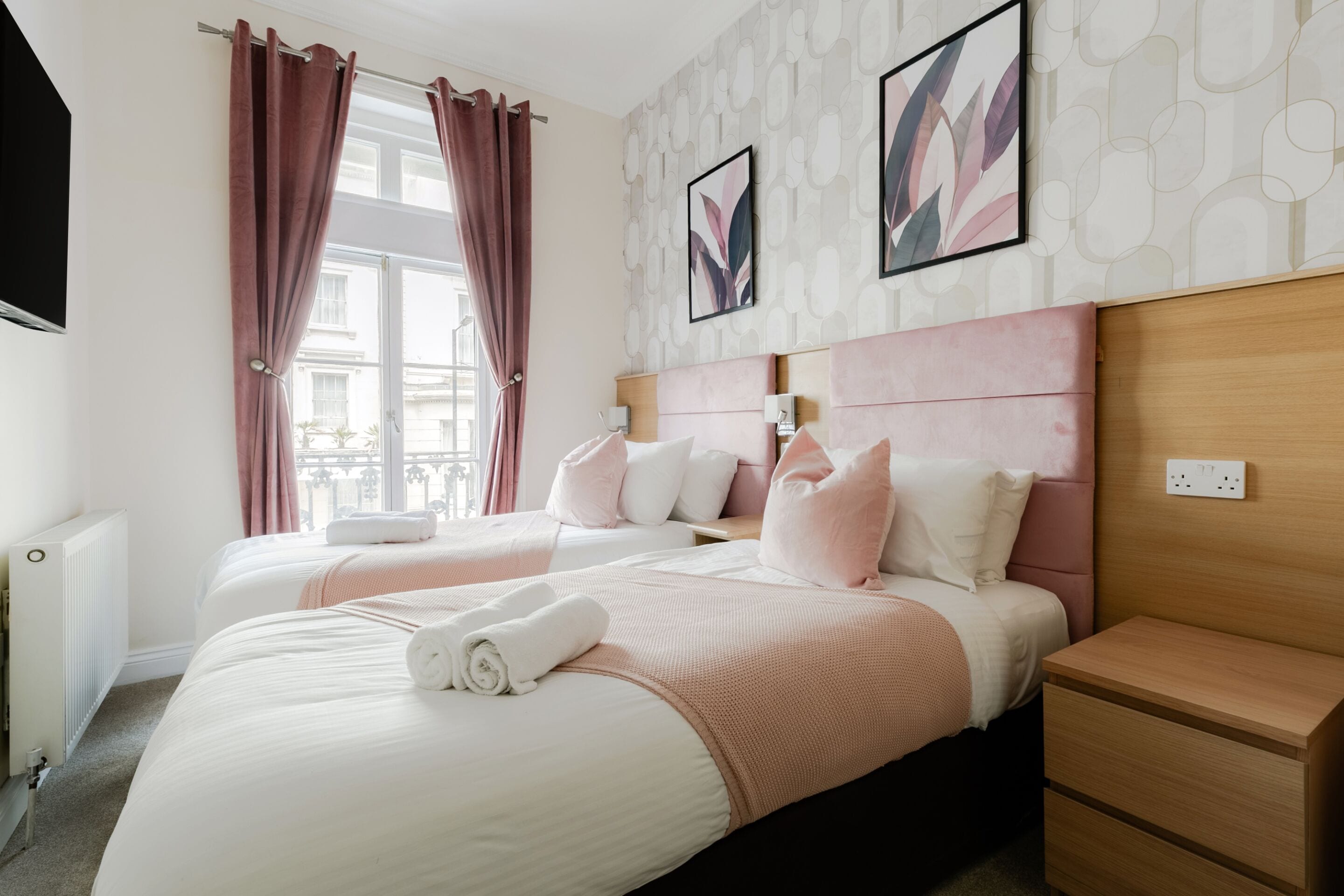 A hotel room with two beds, a wooden headboard, a nightstand, a radiator, and a window with curtains.
