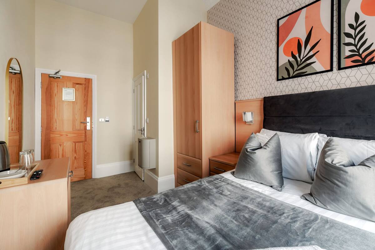 A hotel room with a bed, bedside table, wardrobe, and a wall with artwork.