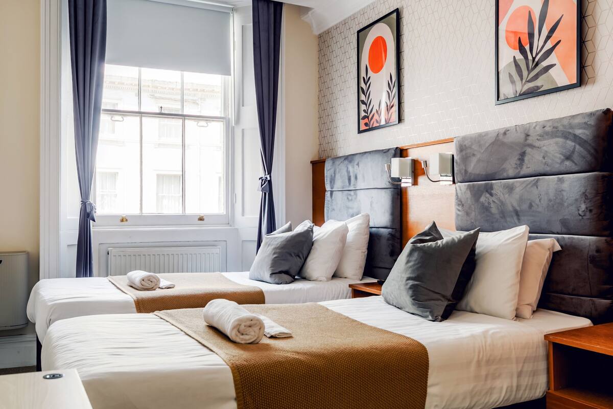 A hotel room with two beds, a large window with curtains, a headboard, and artwork on the wall.
