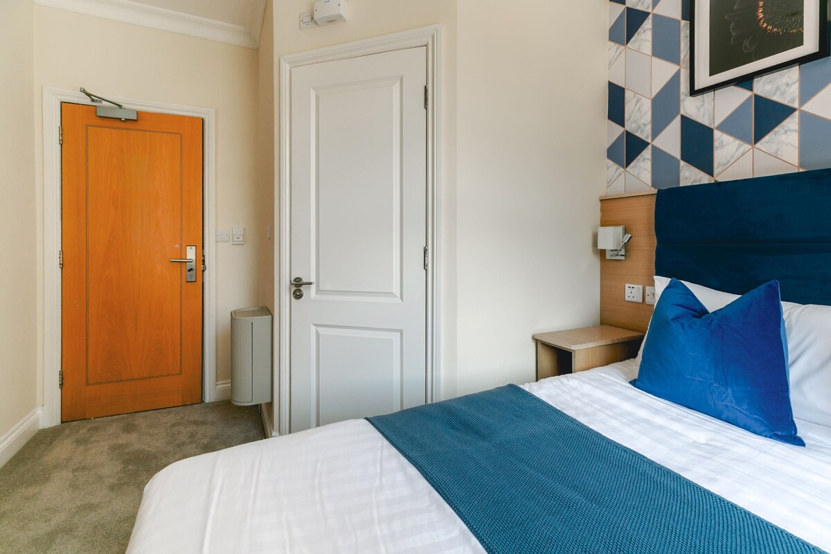 A hotel room with a bed, a wooden door, a white door, a nightstand, a blue headboard, and a wall with a geometric pattern.