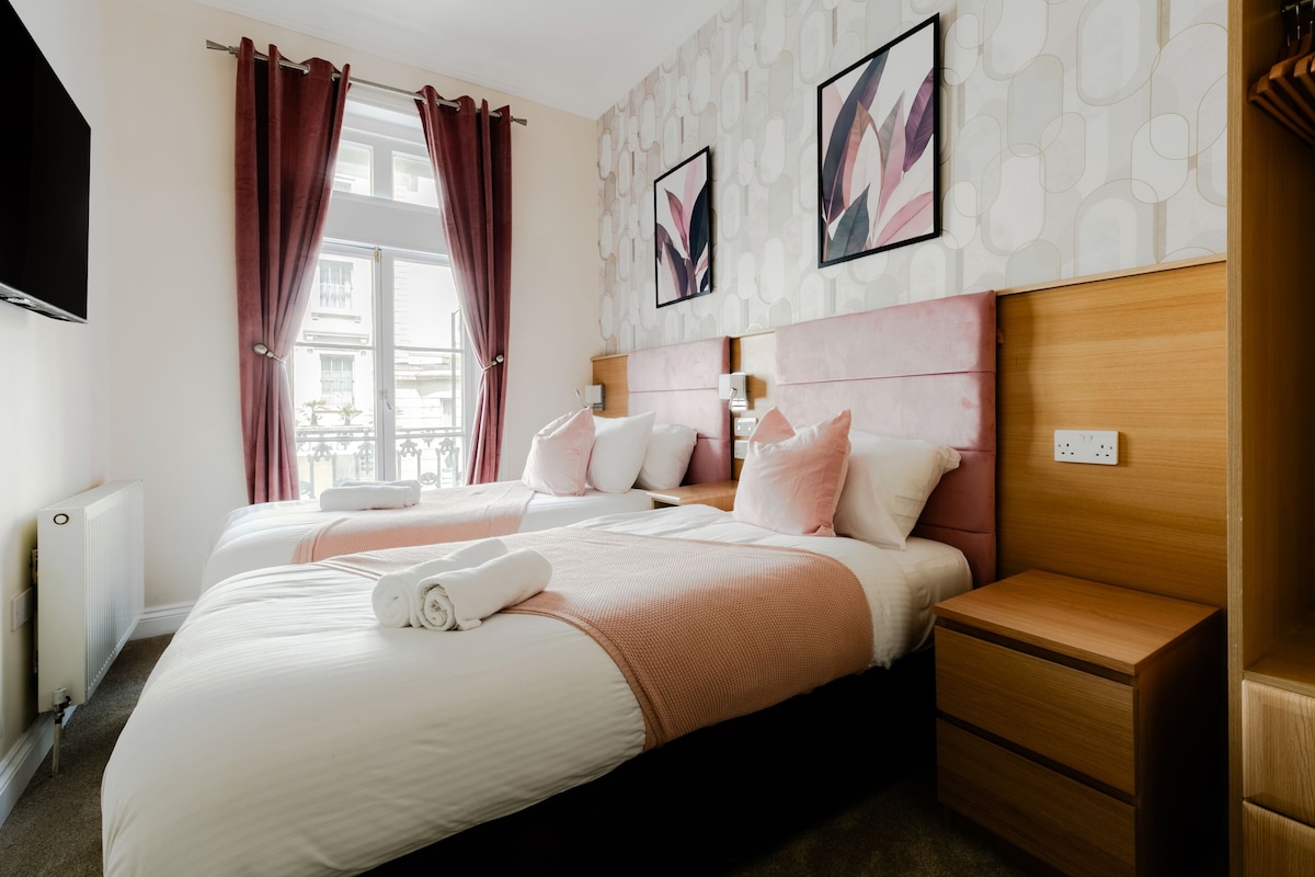 Habitación de hotel con dos camas, cabecero de madera, mesita de noche, televisor montado en la pared y ventana con cortinas.