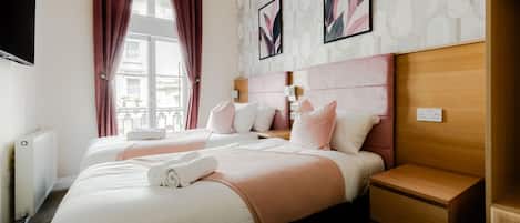 A hotel room with two beds, a wooden headboard, a nightstand, a wall-mounted TV, and a window with curtains.