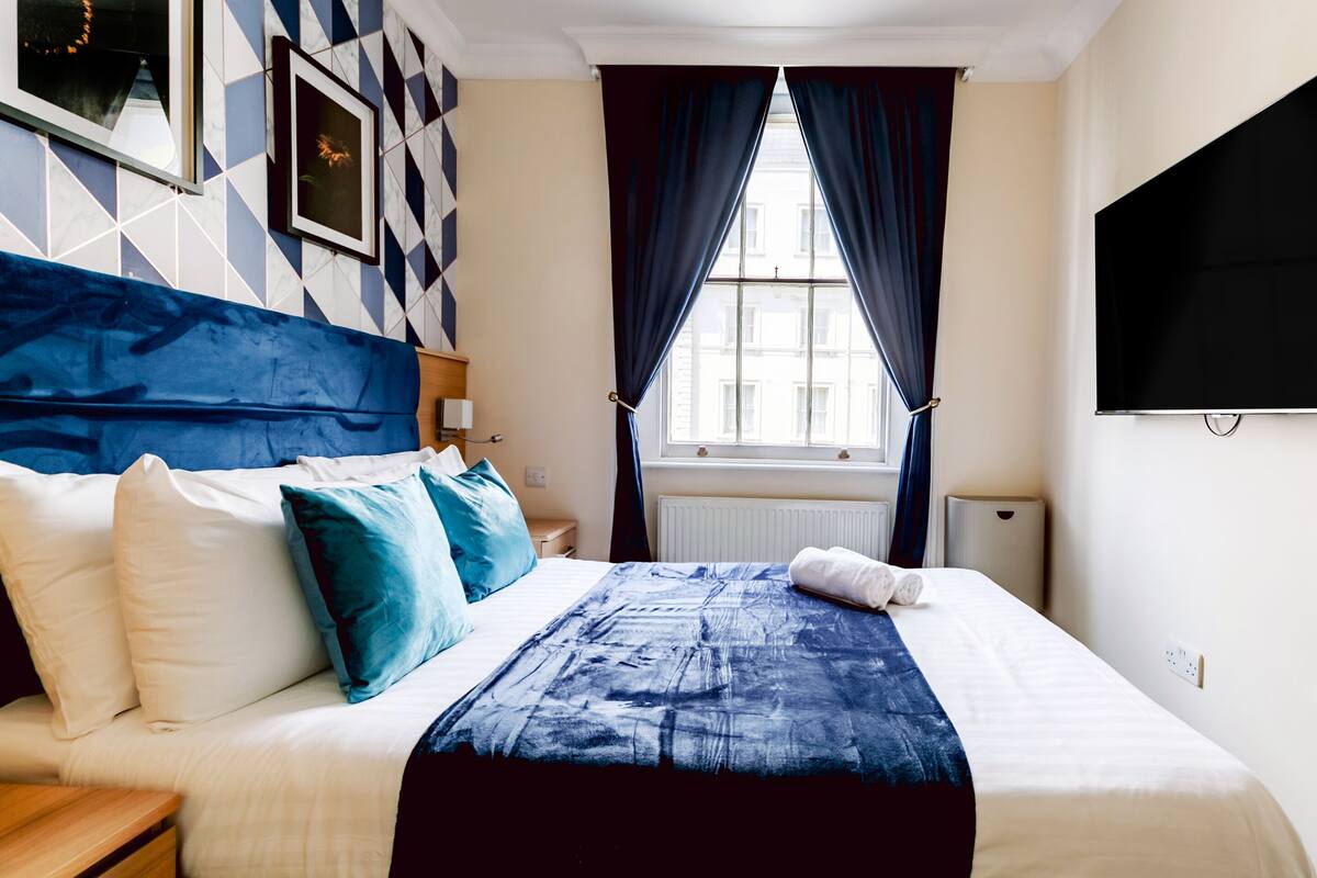 A hotel room with a bed, a TV, a window with curtains, and a geometric wall design.