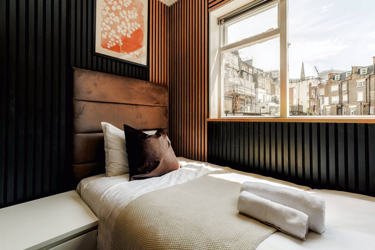 A bedroom with a wooden headboard, a window with a city view, a bed with a white comforter, and a brown pillow.