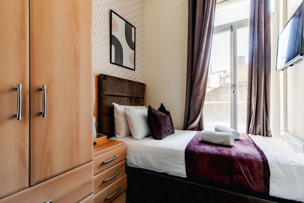 A hotel room with a wooden wardrobe, a bed with a red blanket, a headboard, and a window with curtains.