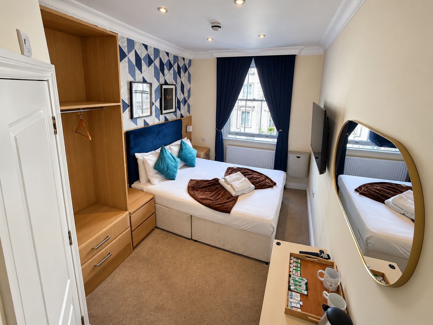 A hotel room with a bed, a wardrobe, a desk with a mirror, and a window with curtains.