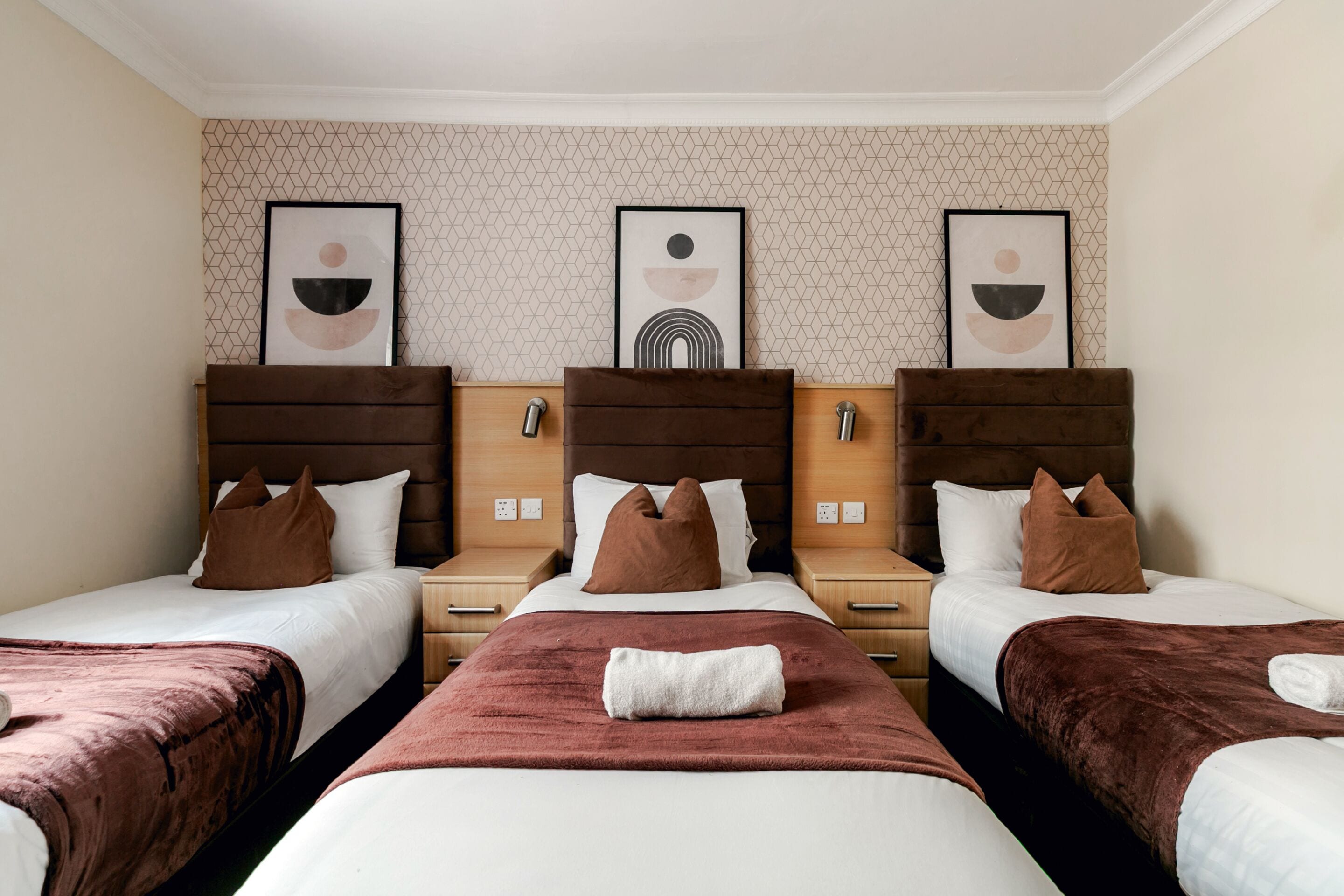 A hotel room with three beds, each with a brown headboard, white bedding, and a brown throw blanket. There are framed pictures on the wall above the beds.