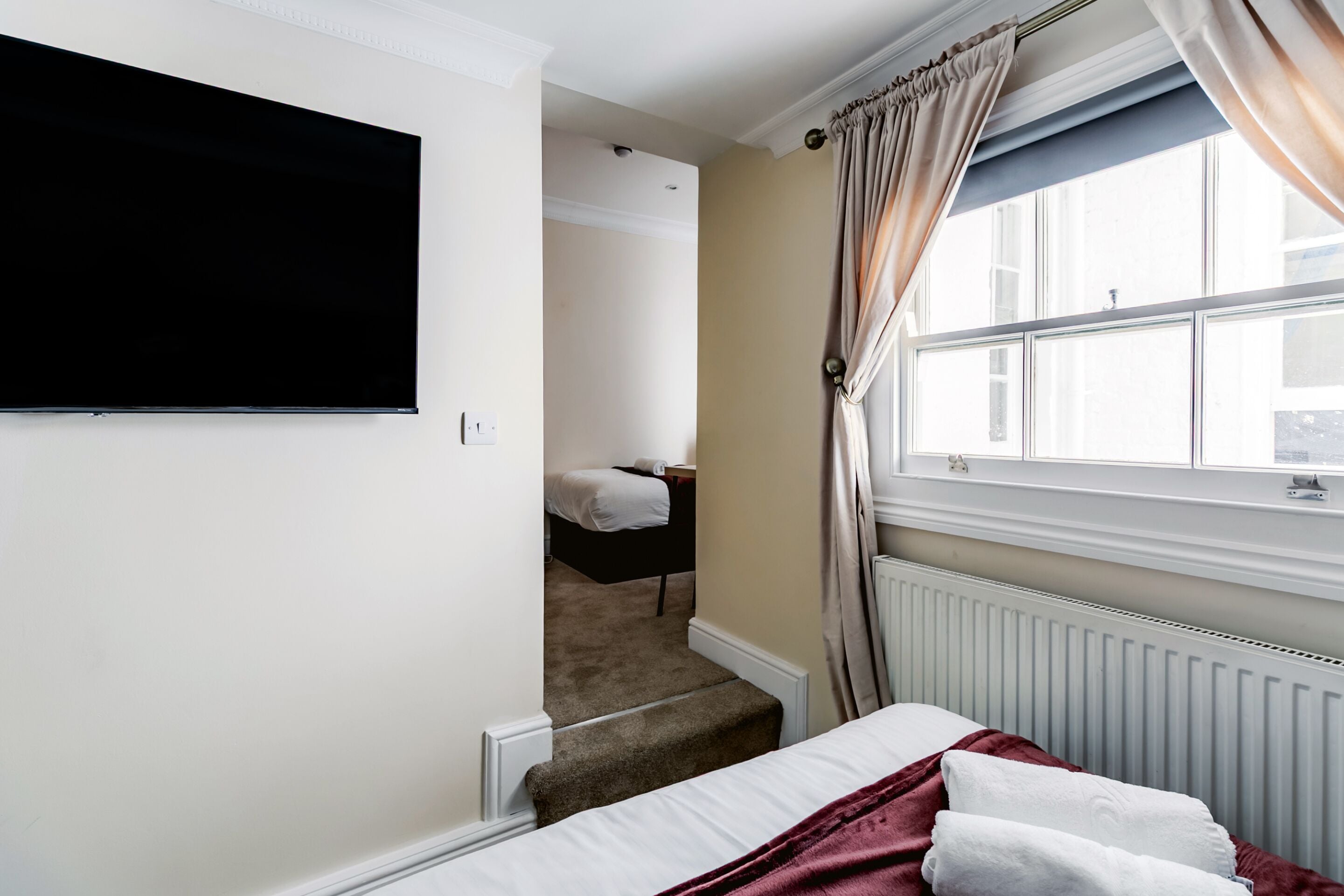 A hotel room with a bed, a TV mounted on the wall, a window with curtains, and a radiator.
