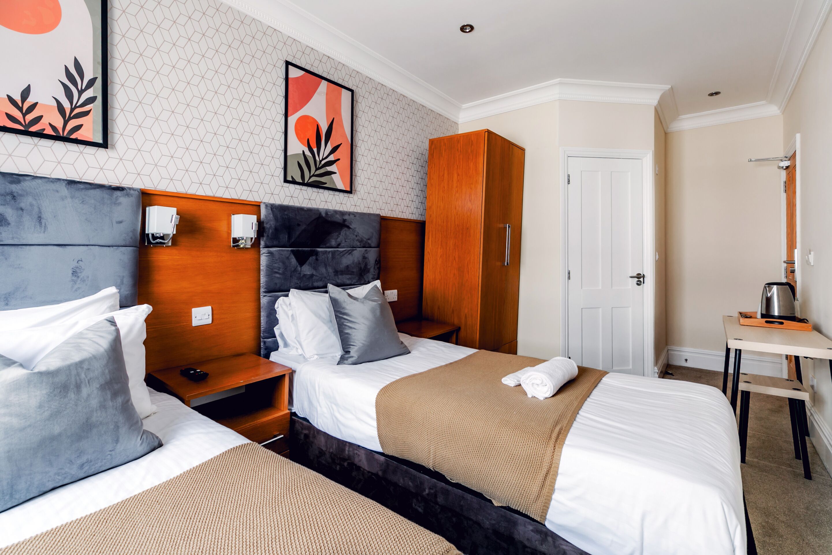 A hotel room with two beds, a wardrobe, a desk with a kettle, and artwork on the walls.