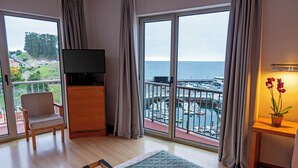A room with a bed, a TV, a chair, a small table with a plant, and a view of the sea through large windows.
