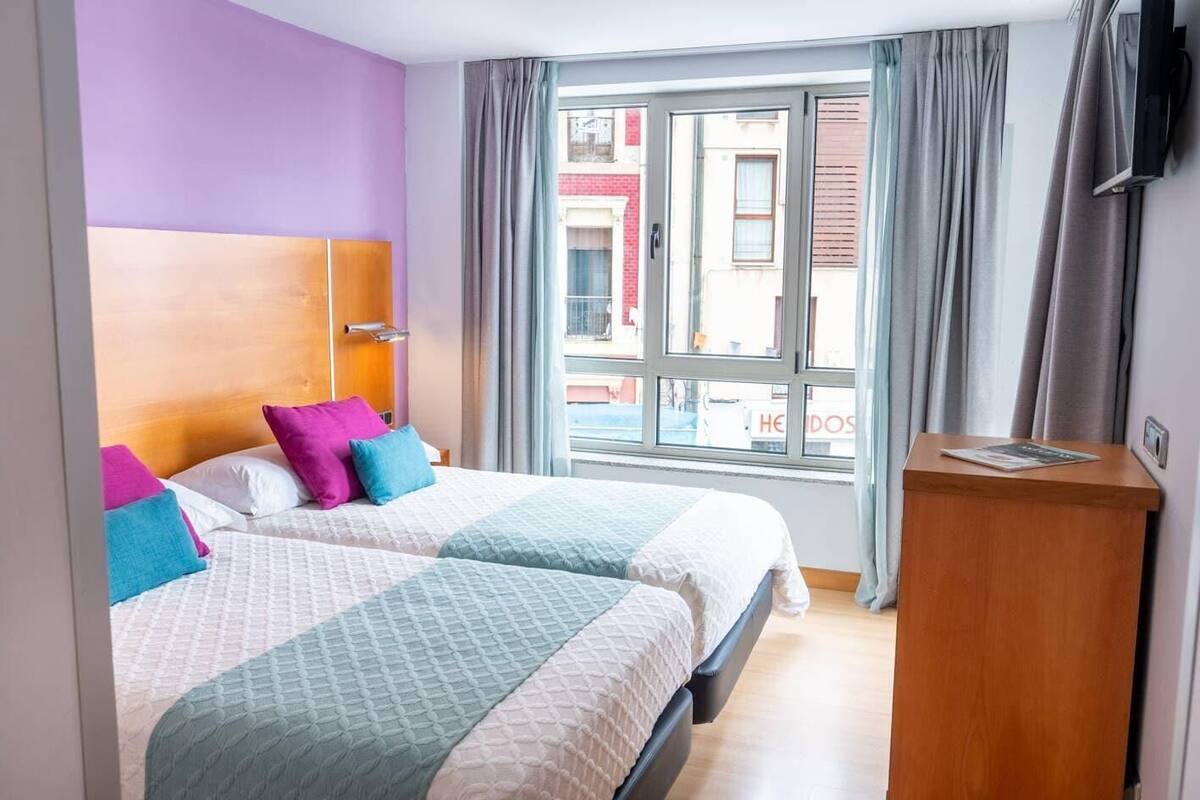 Een hotelkamer met twee bedden, een houten hoofdbord, een bureau van hout en een raam met gordijnen.