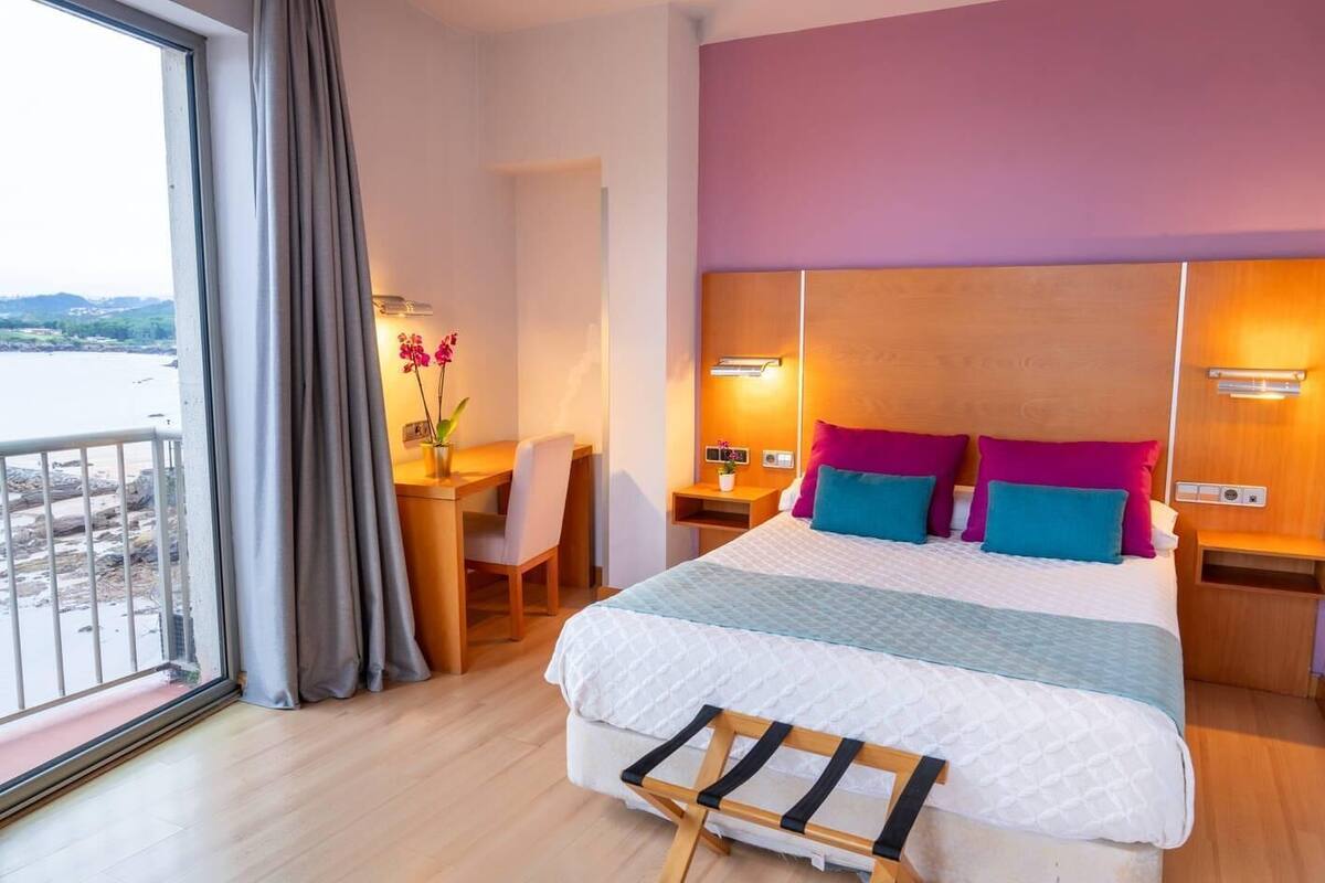 Een hotelkamer met een groot bed, een bureau en een stoel. Door het raam is er uitzicht op het strand.