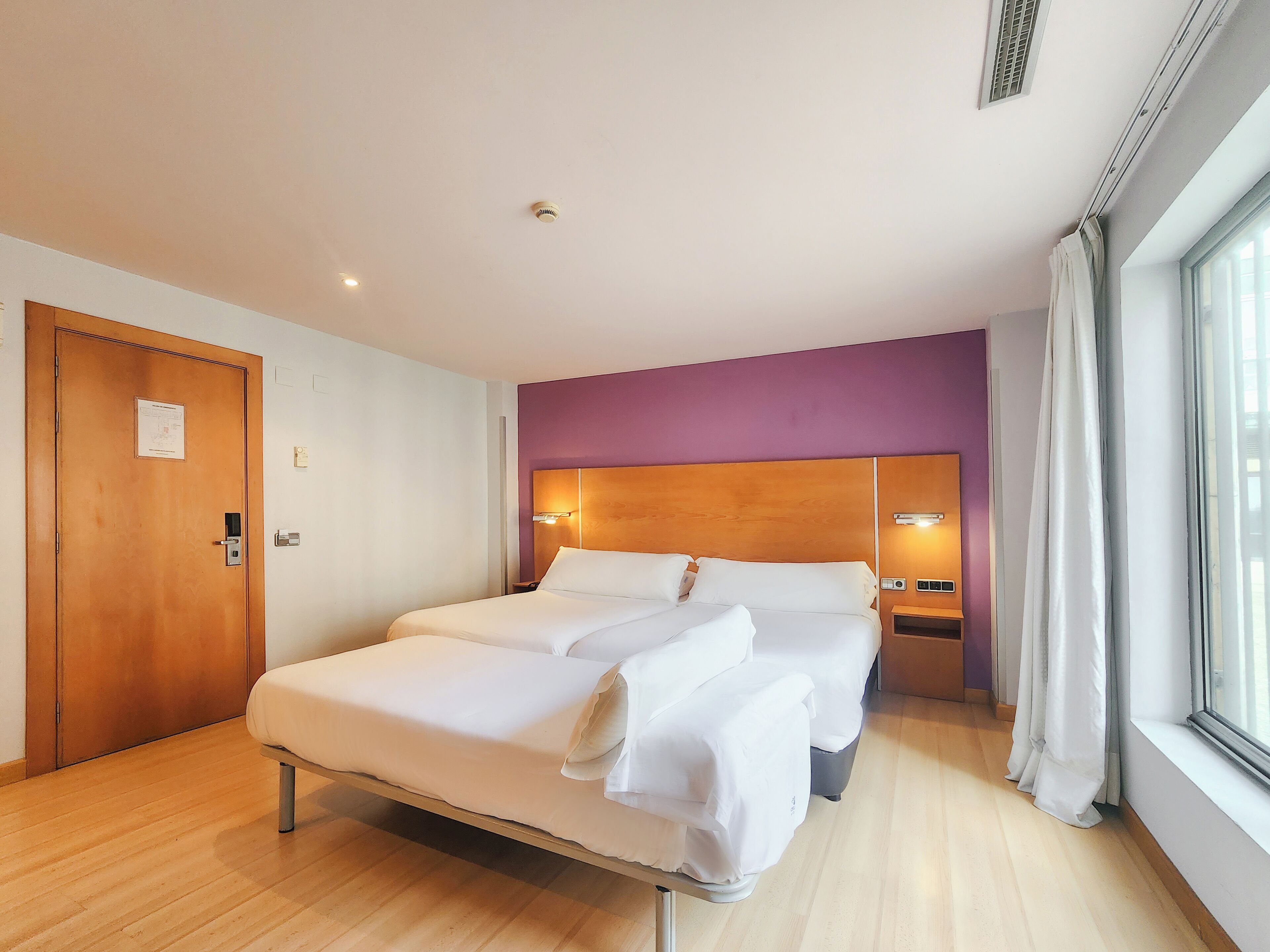 Habitación de hotel con una cama grande, una puerta de madera, una pared de acento púrpura y una ventana con cortinas.